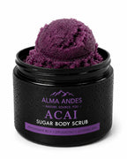 A black jar of Alma Andes Acai Sugar Body Scrub, shown open with a scoop of deep purple scrub visible inside. The jar has a textured black finish with condensation droplets, and the label features purple and white text.