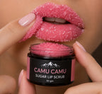 A close-up of a person holding a jar of sugar lip scrub while applying a sparkling pink lip scrub to their lips. The jar is labeled 'CAMU CAMU SUGAR LIP SCRUB 50 gm' by the brand 'LUMA ANDEI'.