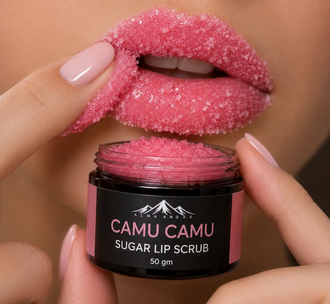 A close-up of a person holding a jar of sugar lip scrub while applying a sparkling pink lip scrub to their lips. The jar is labeled 'CAMU CAMU SUGAR LIP SCRUB 50 gm' by the brand 'LUMA ANDEI'.