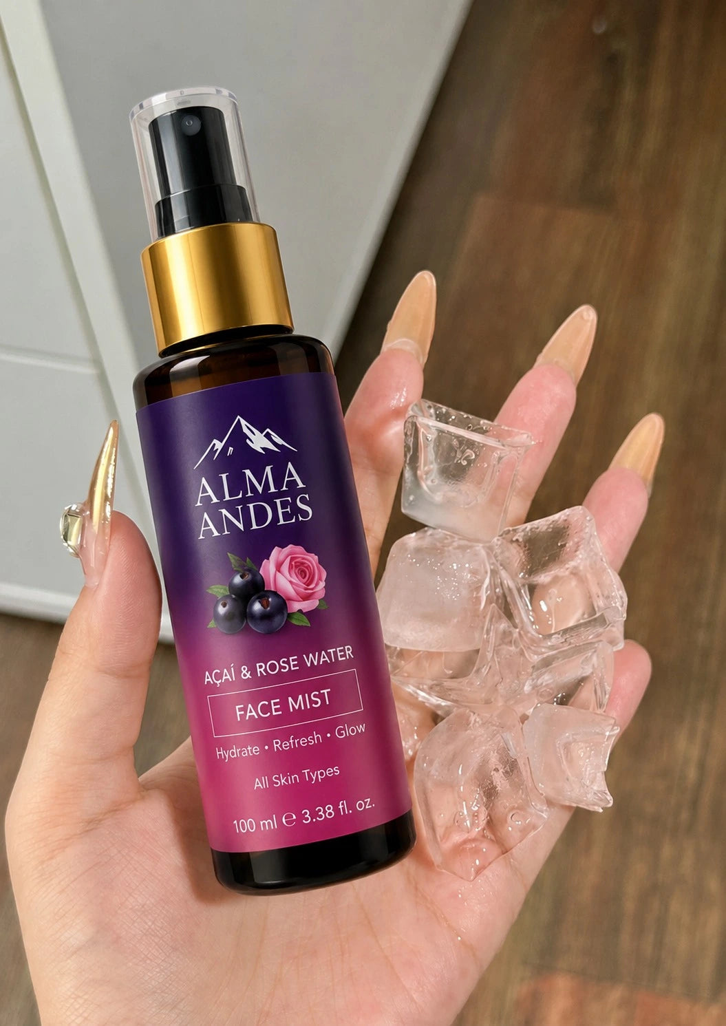 A hand holding a bottle of Almaandes Acai & Rose Water Face Mist next to several clear ice cubes. The face mist bottle has a dark purple label with a gold pump collar and a black spray top.